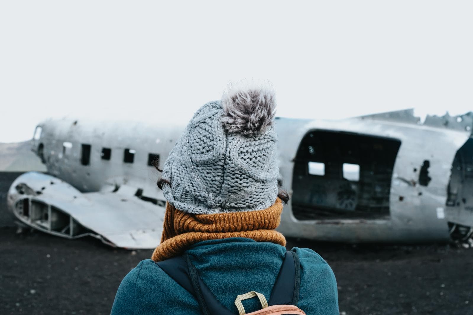 Person mit Strickmütze vor einem verlassenen Flugzeugwrack auf schwarzem Sand in Island.