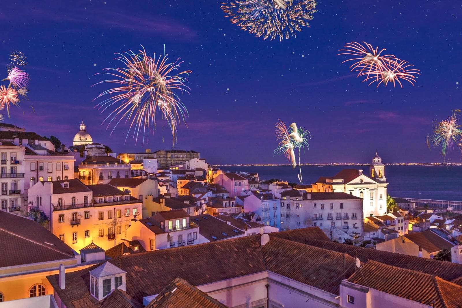 Feuerwerk über Altstadt von Lissabon, Dächer und Meer im Hintergrund, nachtbeleuchtet.