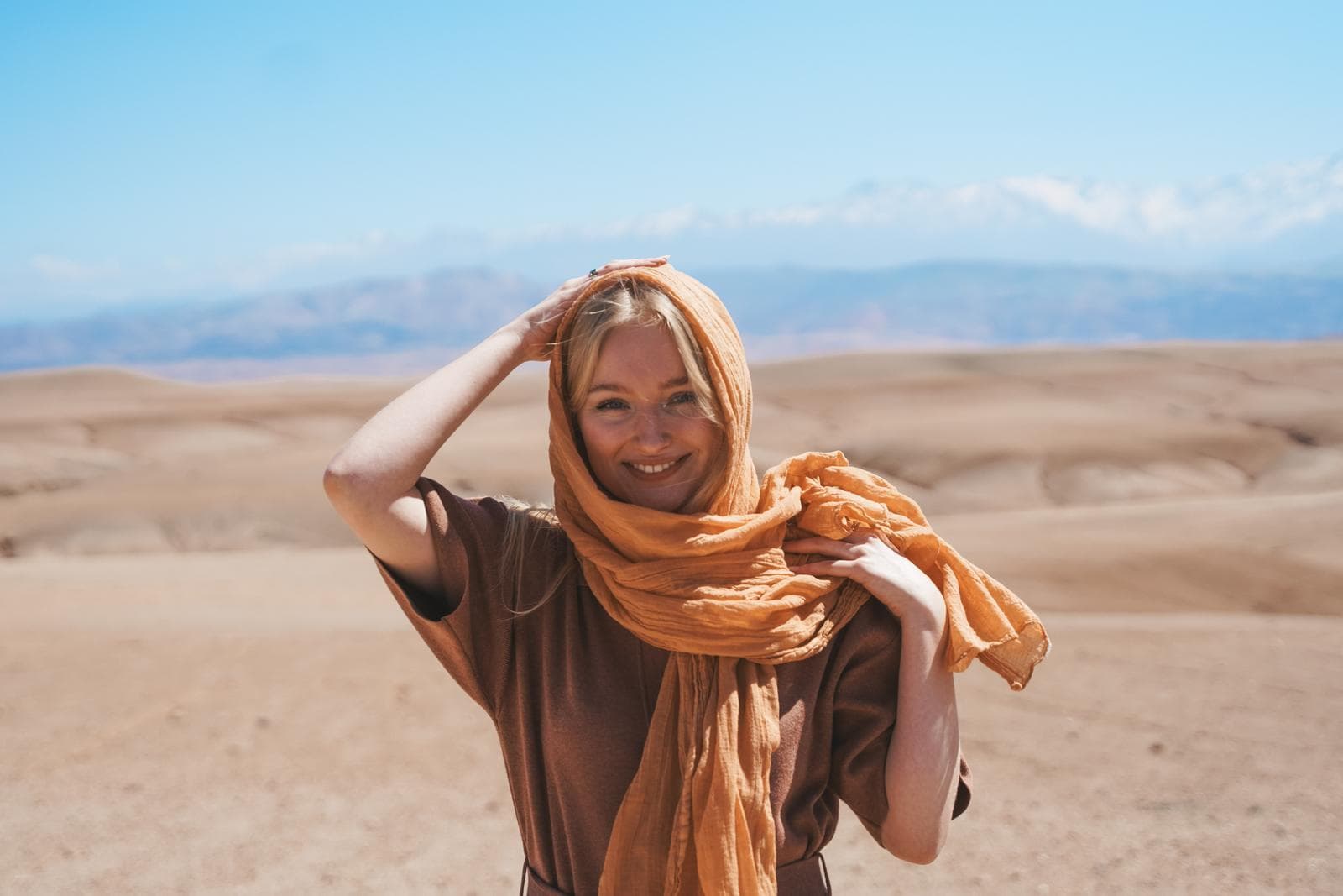 Frau in Wüste mit orangefarbenem Schal, blauer Himmel, sandige Landschaft.