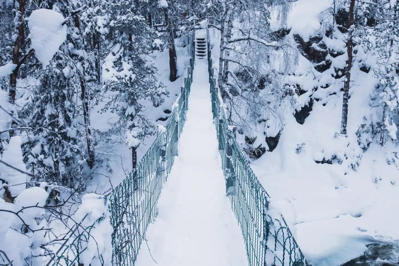Verschneite Hängebrücke in Winterlandschaft, umgeben von schneebedeckten Bäumen.