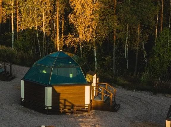 Sechseckige Glashütte im Wald bei Abenddämmerung, umgeben von Herbstbäumen.