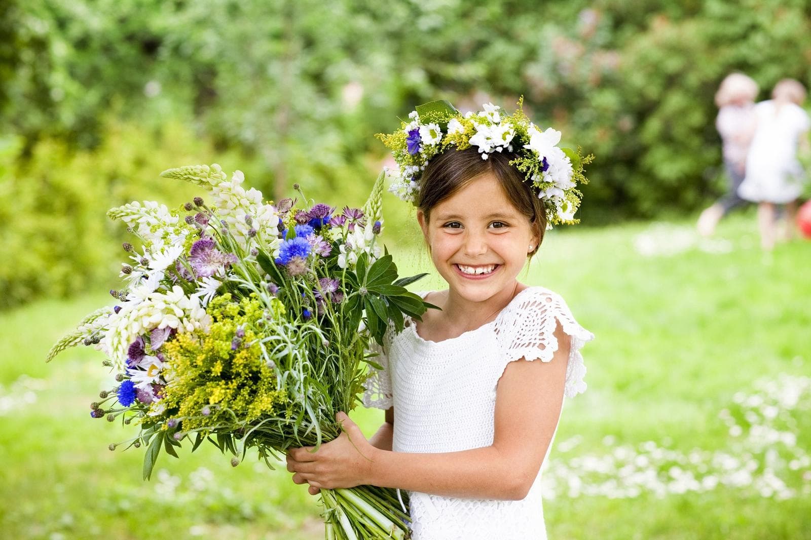 Ein Mädchen in weißem Kleid mit Blumenkranz lächelt, hält bunte Blumen im Garten.