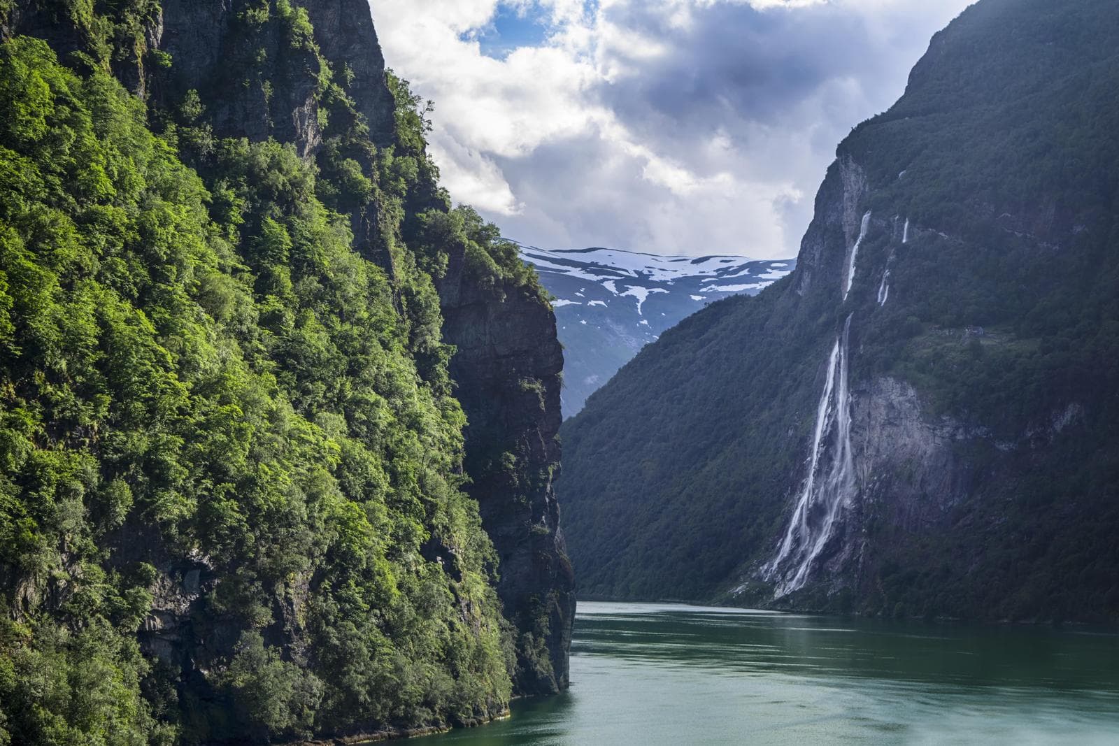 Grüner Fjord in Norwegen mit beeindruckendem Wasserfall, umgeben von steilen, bewaldeten Klippen.