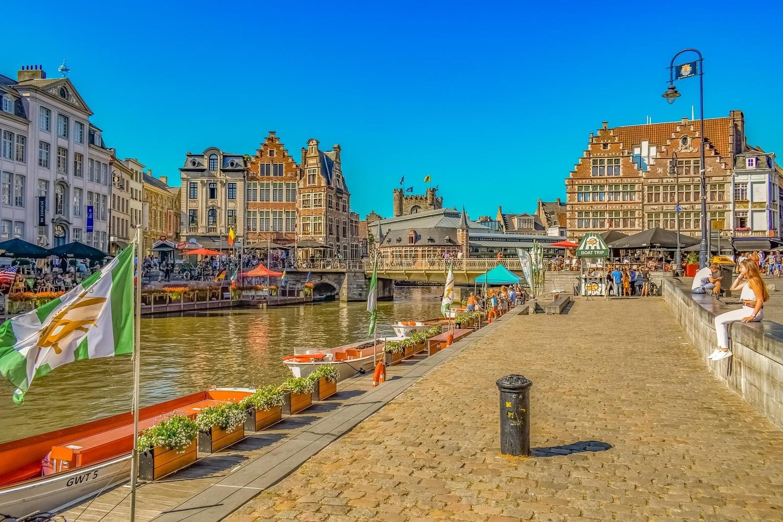 Flussufer in Gent, Belgien mit historischen Gebäuden und Booten bei klarem Himmel.