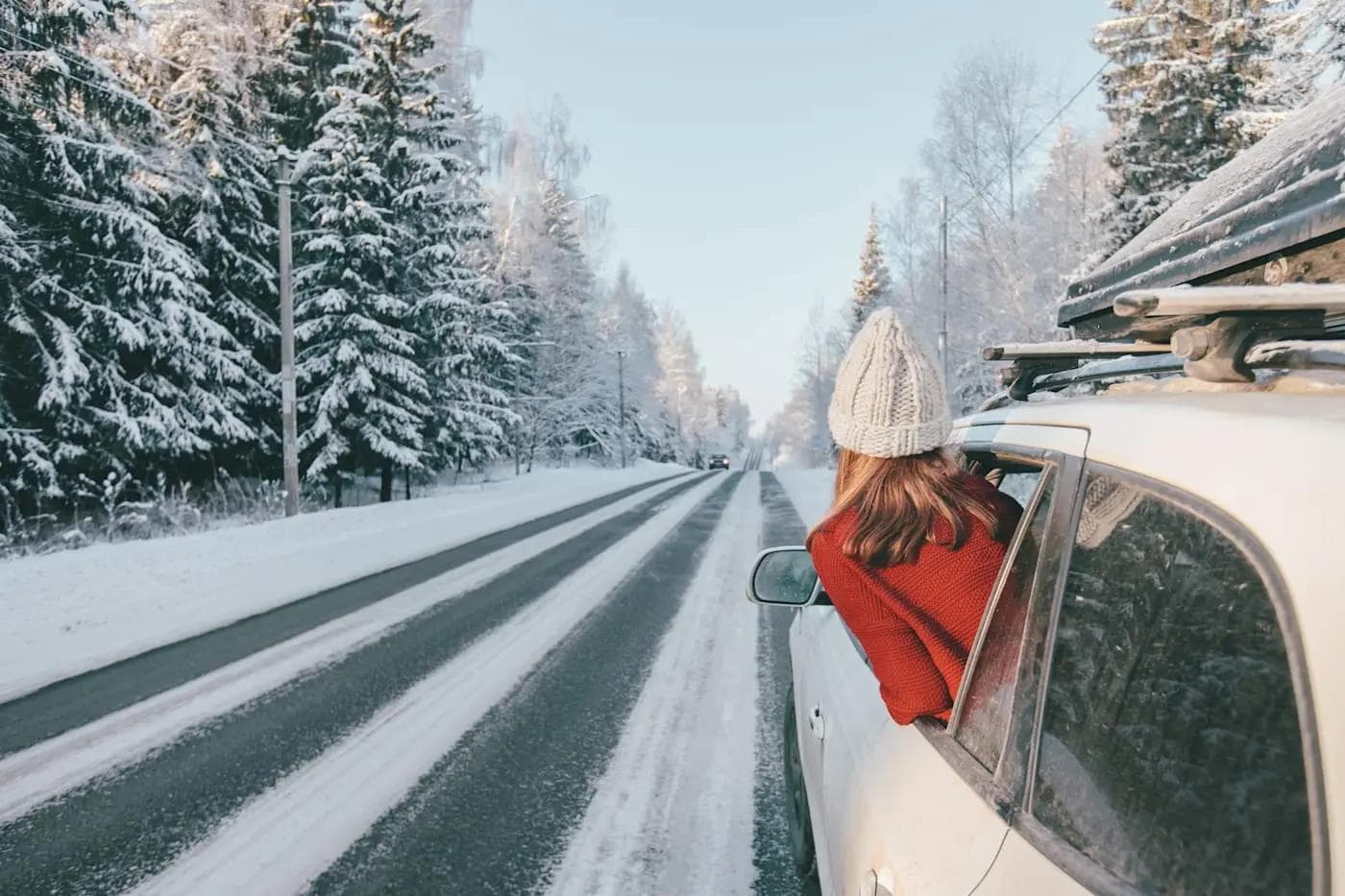 Verschneite Straße durch Winterwald, Frau lehnt sich aus Autofenster, klare Winterluft.