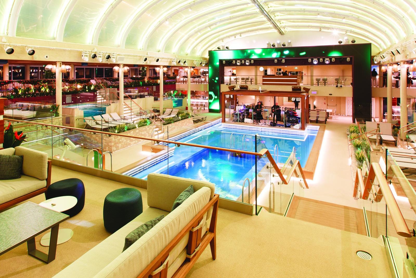 Elegantes Hallenbad mit Glasdecke, stilvollen Sitzbereichen und Live-Bühne.