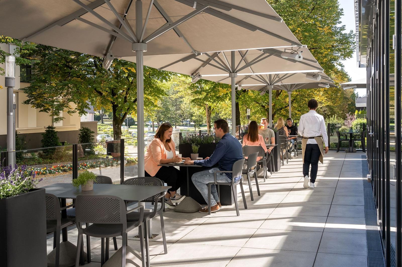 Moderne Hotelterrasse mit Sonnenschirmen, Gästen und Bedienung, umgeben von Bäumen.
