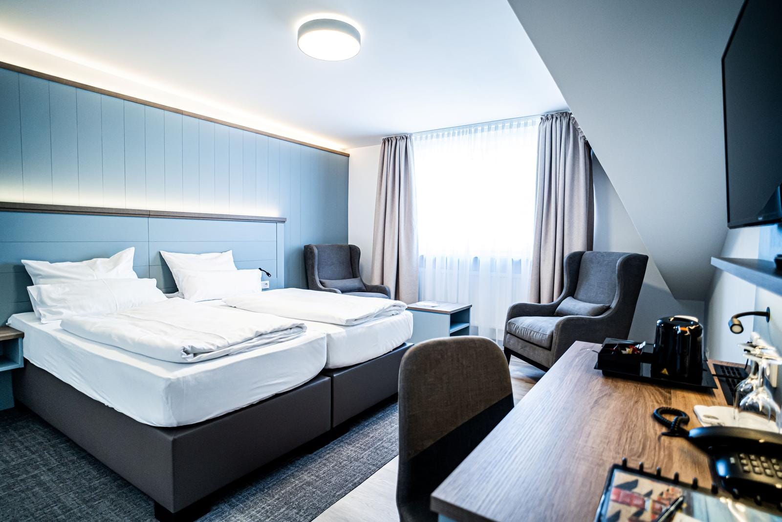 Modernes Hotelzimmer mit zwei Einzelbetten, grauen Sesseln und Holzschreibtisch.