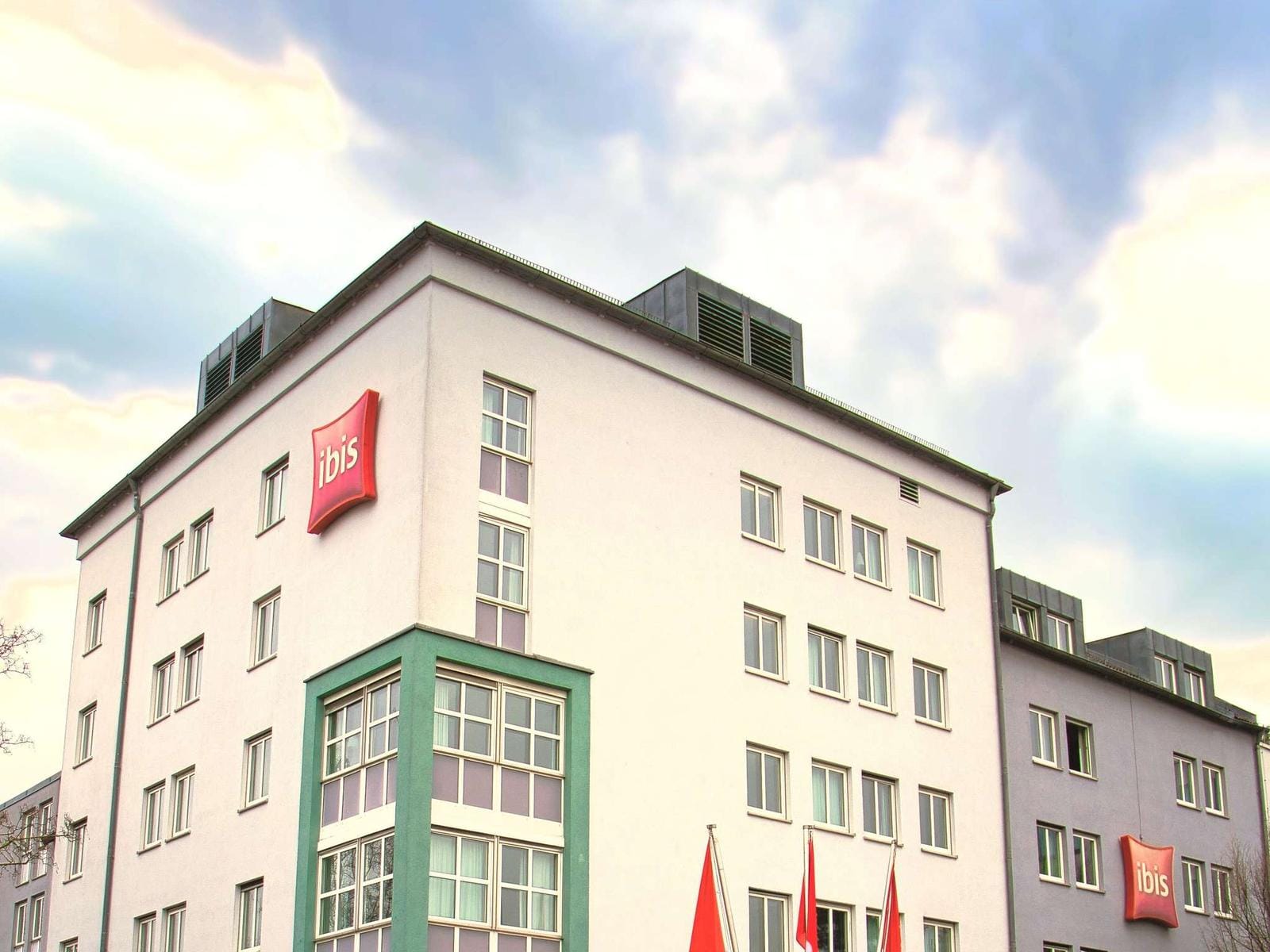 Moderner Hotelbau mit heller Fassade und Ibis-Logo, vor bewölktem Himmel.