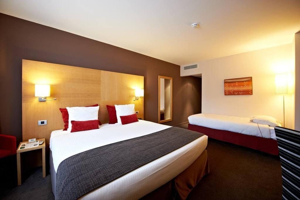 Modernes Hotelzimmer mit Doppel- und Einzelbett, roten Akzenten und stimmungsvollem Licht.