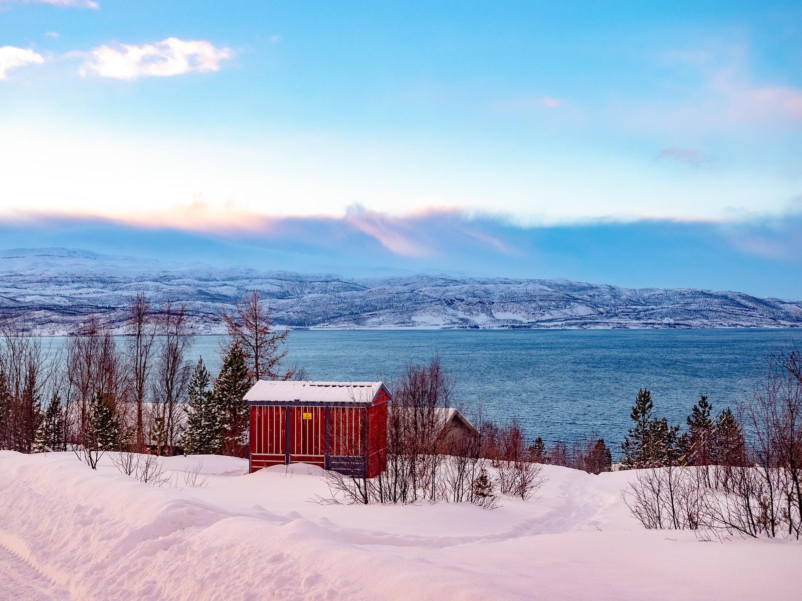 Winterliche norwegische Landschaft mit rotem Haus am See, schneebedeckten Bäumen und Bergen.