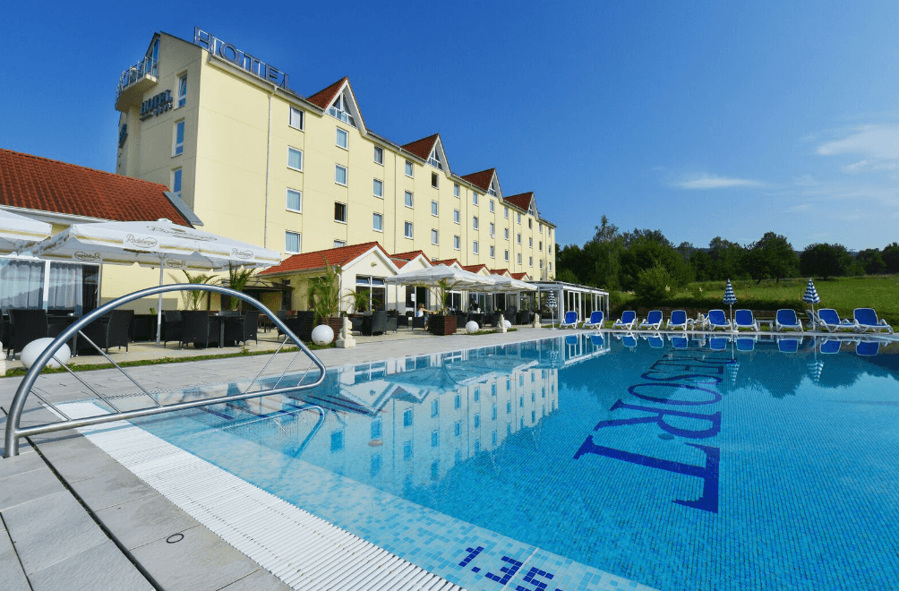Hotel mit blauem Pool, Liegestühlen, gelbem Gebäude, grüner Umgebung unter blauem Himmel.
