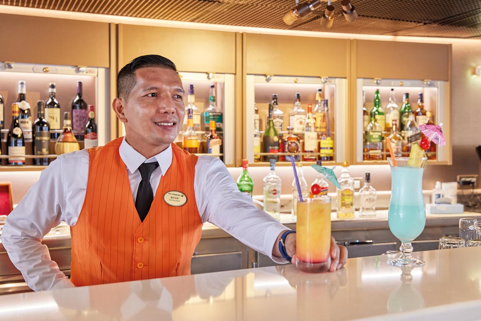 Barmann in orangefarbener Weste serviert bunte Cocktails in eleganter Hotelbar.