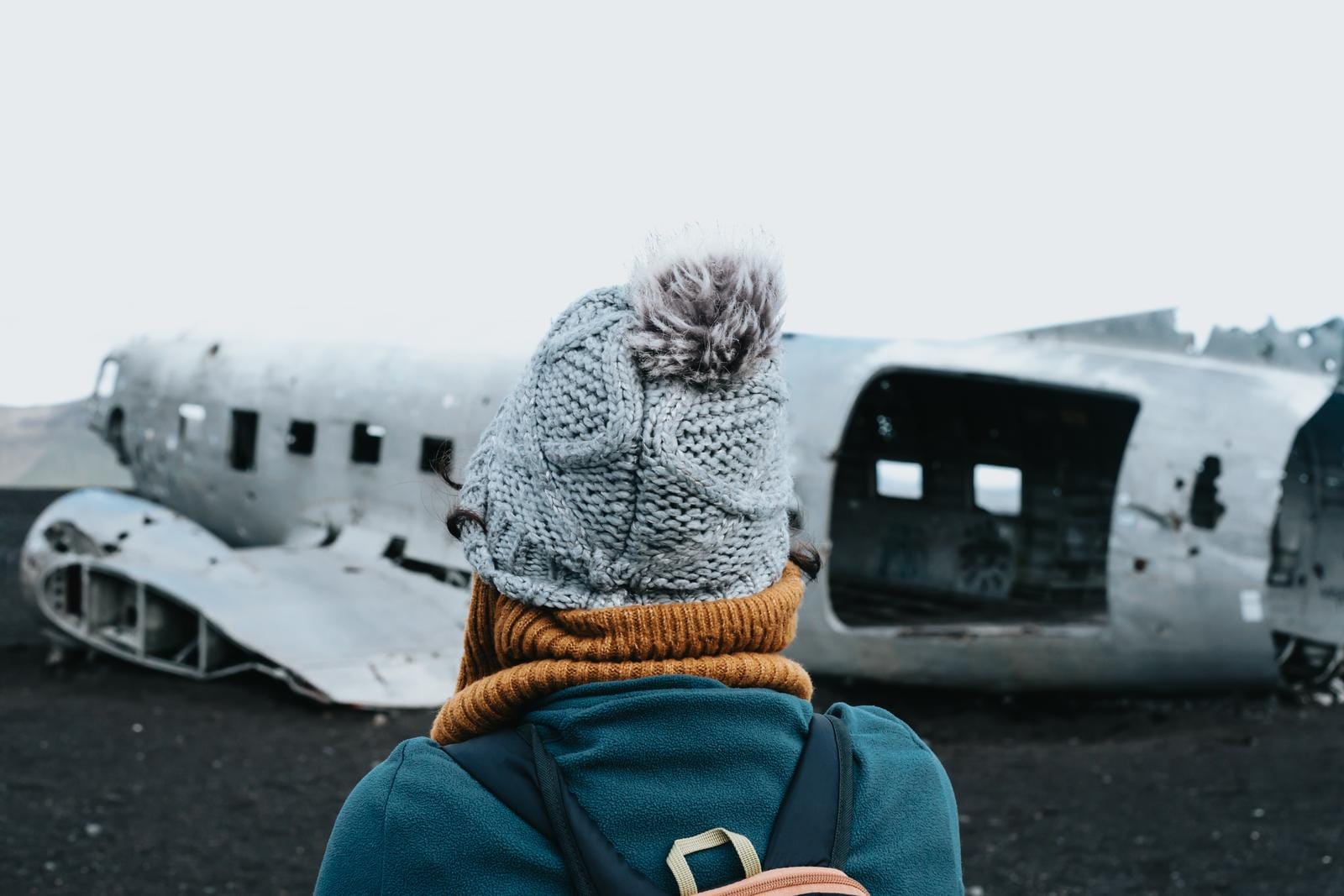 Person mit Strickmütze vor einem verlassenen Flugzeugwrack auf schwarzem Sand in Island.