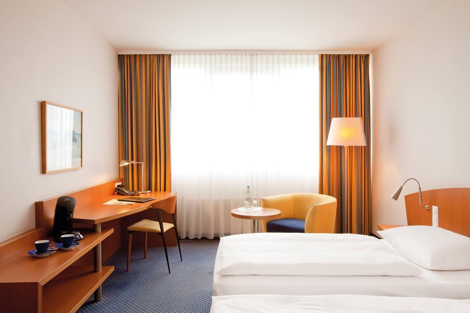 Helles Hotelzimmer mit modernem Holzschreibtisch, gelbem Sessel und gestreiften Vorhängen.