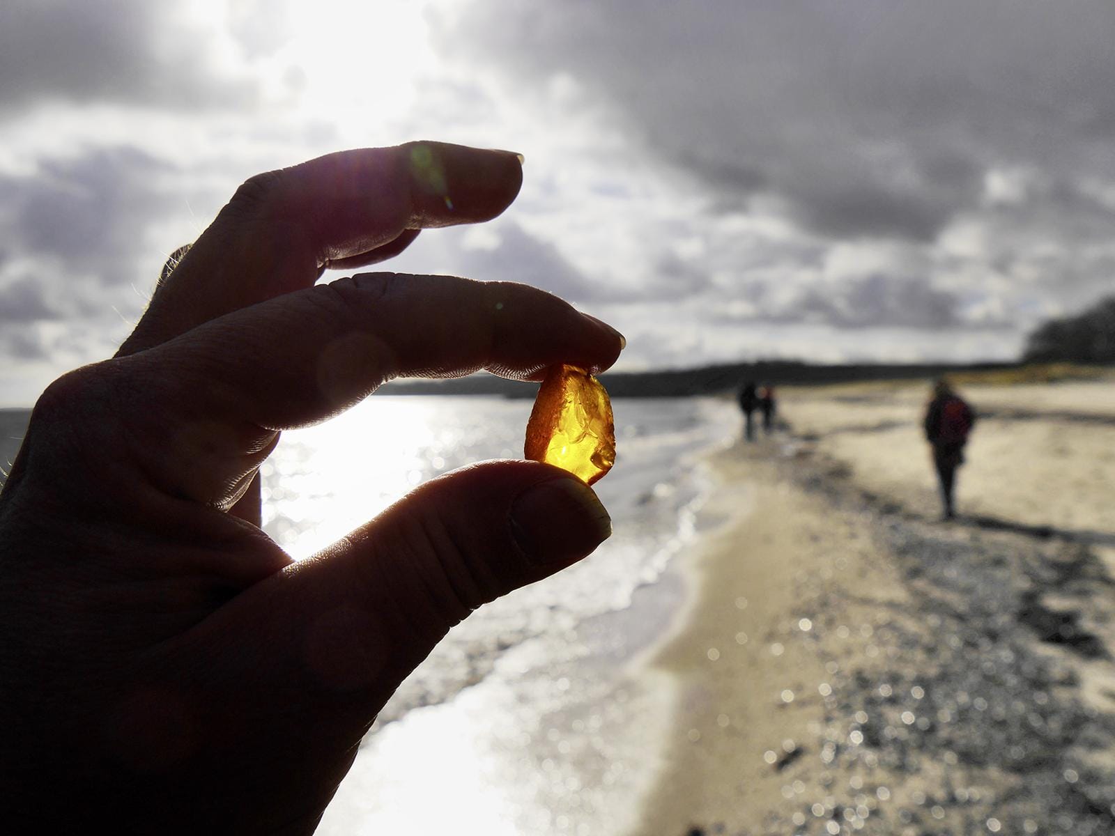 Person hält Bernstein am sonnigen Strand von Rügen, mit Spaziergängern im Hintergrund.
