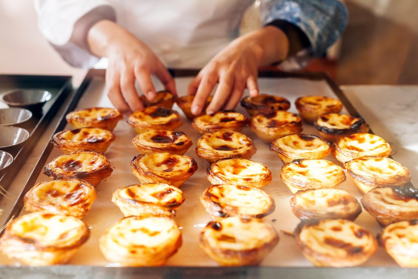 Hände arrangieren goldene Pastéis de Nata auf Backblech in warmem Licht.