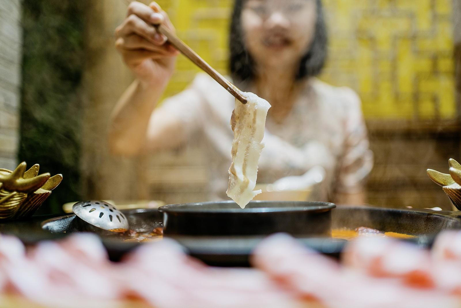 Person genießt Hotpot in einem Restaurant mit dampfenden Brühen und gelber Wand.