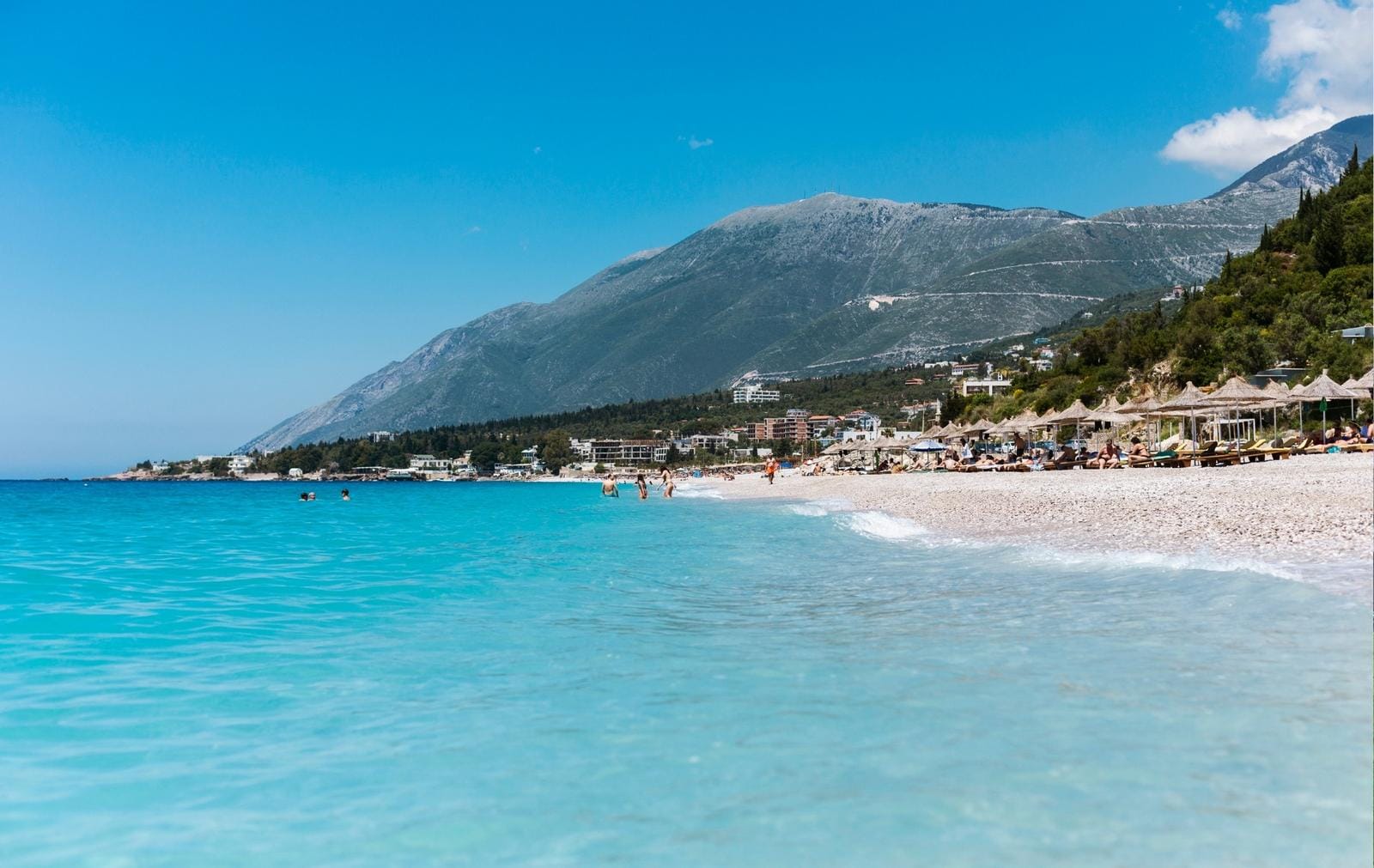 Klarer Strand in Albanien mit türkisfarbenem Meer, Bergen und Sonnenschirmen.