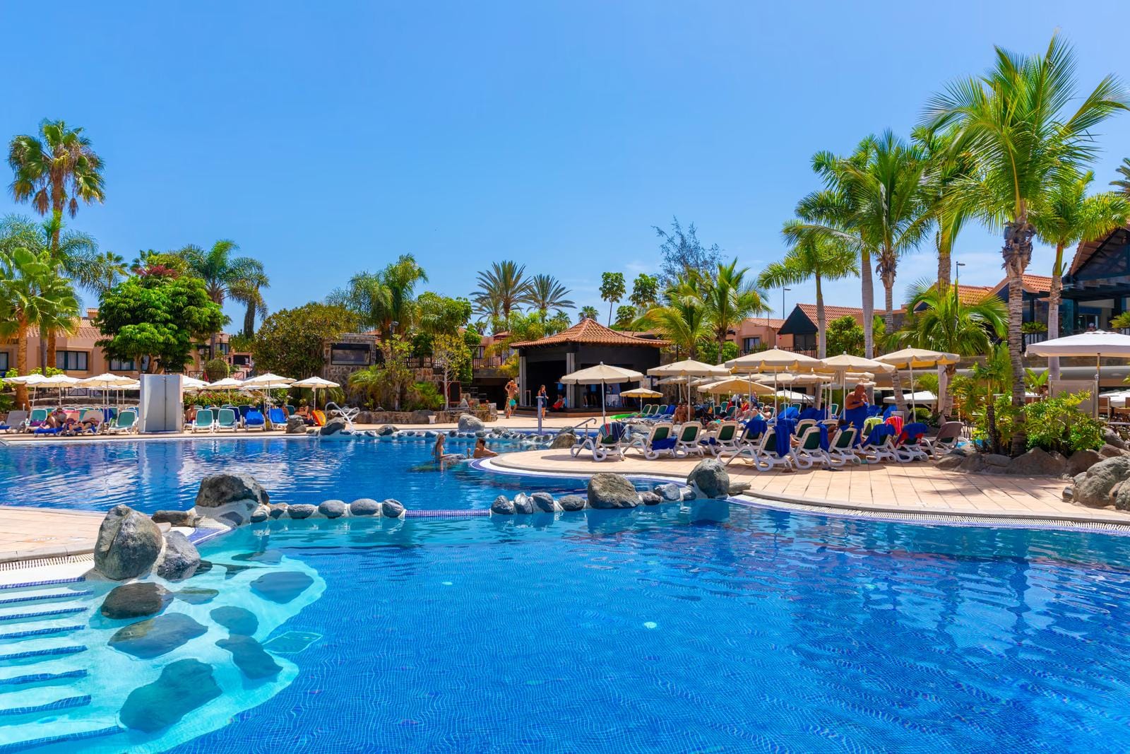 Großer Hotelpool mit Palmen, Sonnenliegen und Sonnenschirmen unter blauem Himmel.