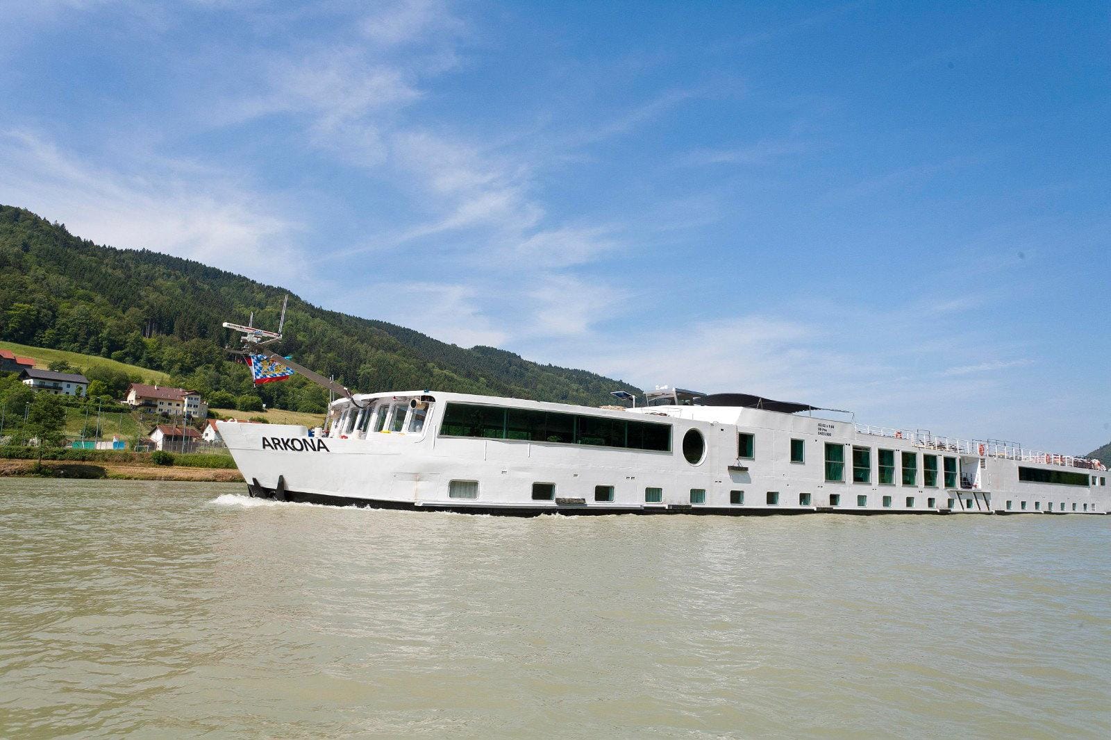 Weißes Flusskreuzfahrtschiff auf einem Fluss vor grünen Hügeln und blauem Himmel.