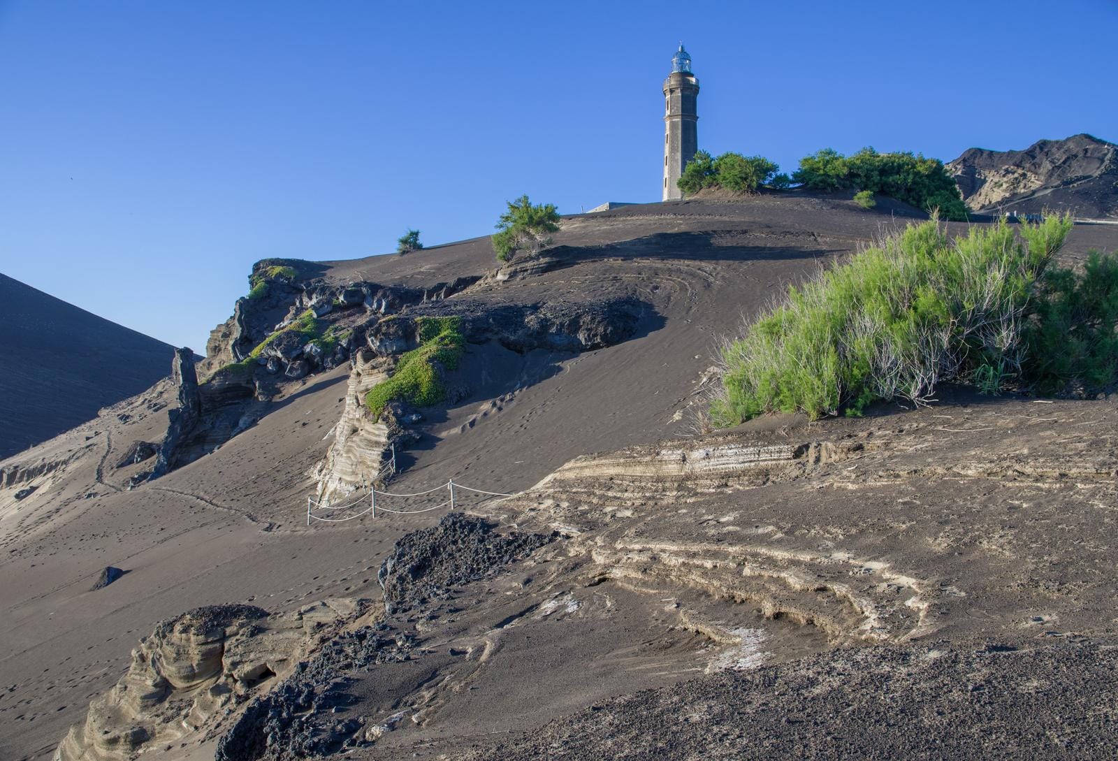 Vulkanlandschaft mit Leuchtturm und schwarzem Sand unter blauem Himmel.