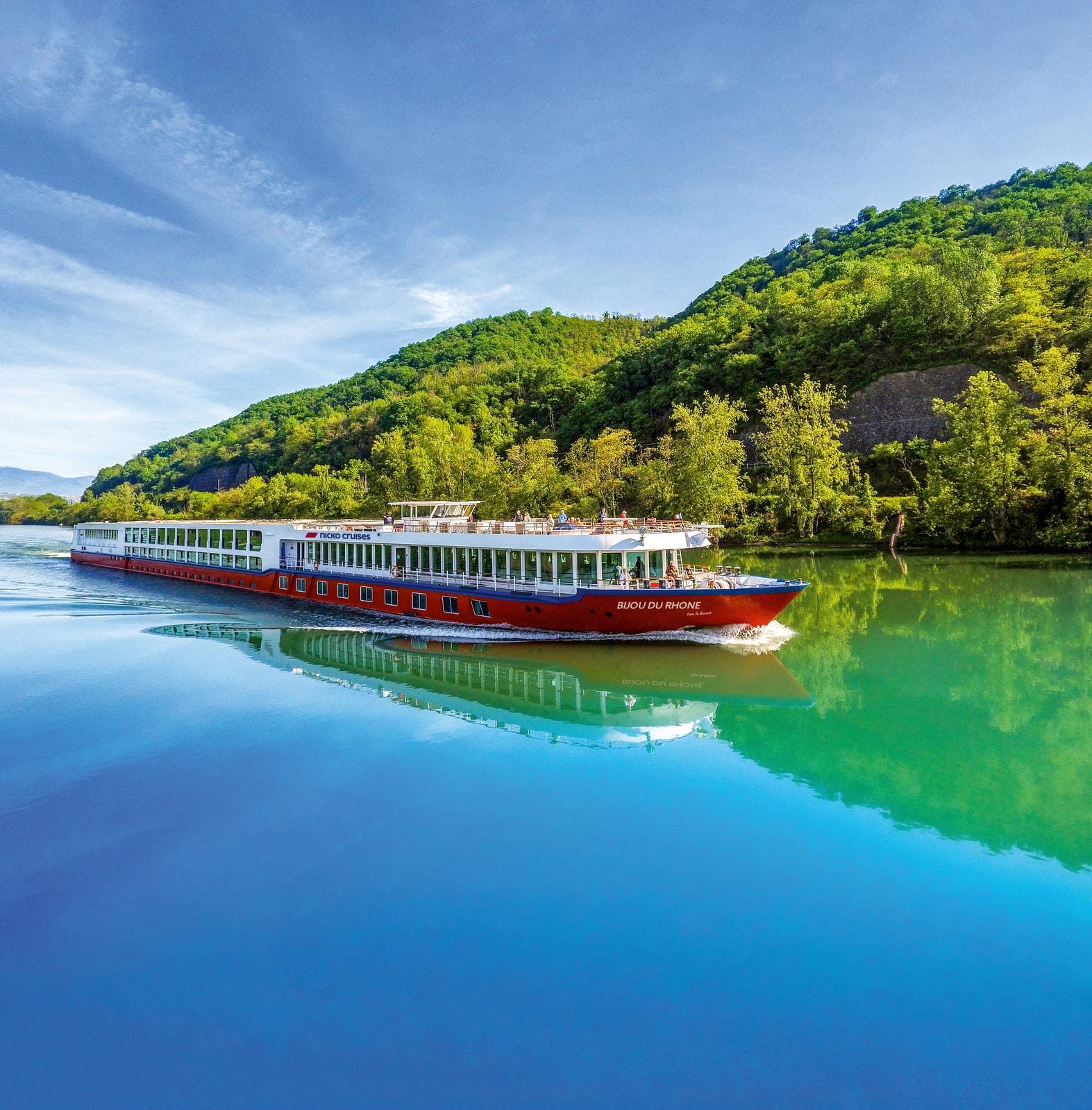 Bijou du Rhone auf grünem Fluss, umgeben von grünen Hügeln unter blauem Himmel.