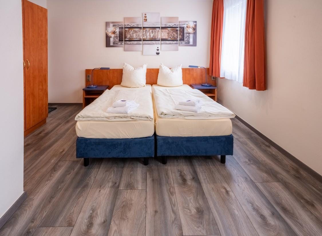 Helles Hotelzimmer mit Doppelbett, Holzmöbeln, Teppich, Kunstbild und orangenen Vorhängen.