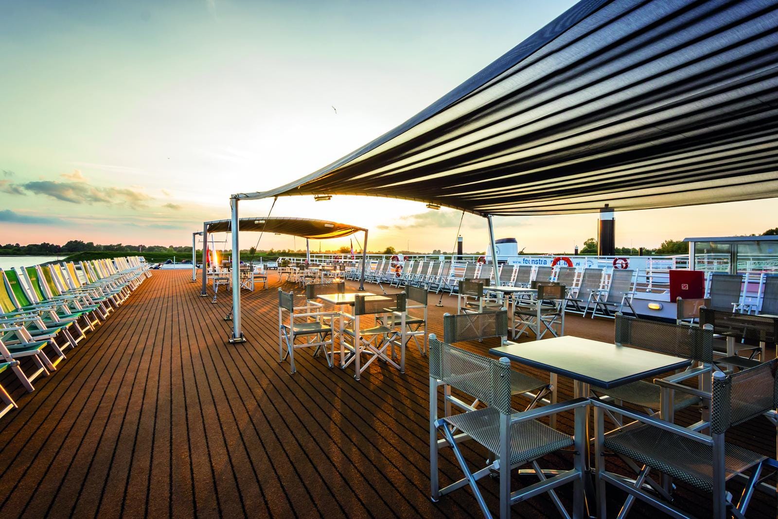 Sonnendeck eines Kreuzfahrtschiffs bei Sonnenuntergang, mit Liegestühlen und Tischen.