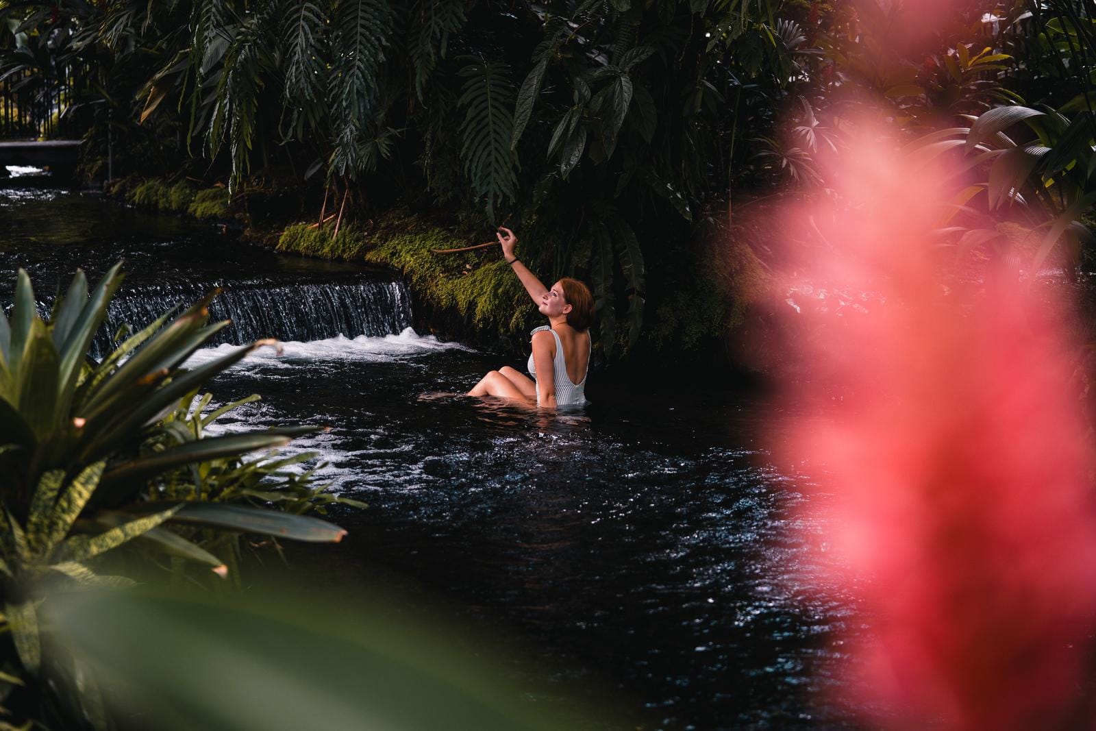 Frau im weißen Badeanzug sitzt im tropischen Wasserfall-Pool, umgeben von üppigem Grün.
