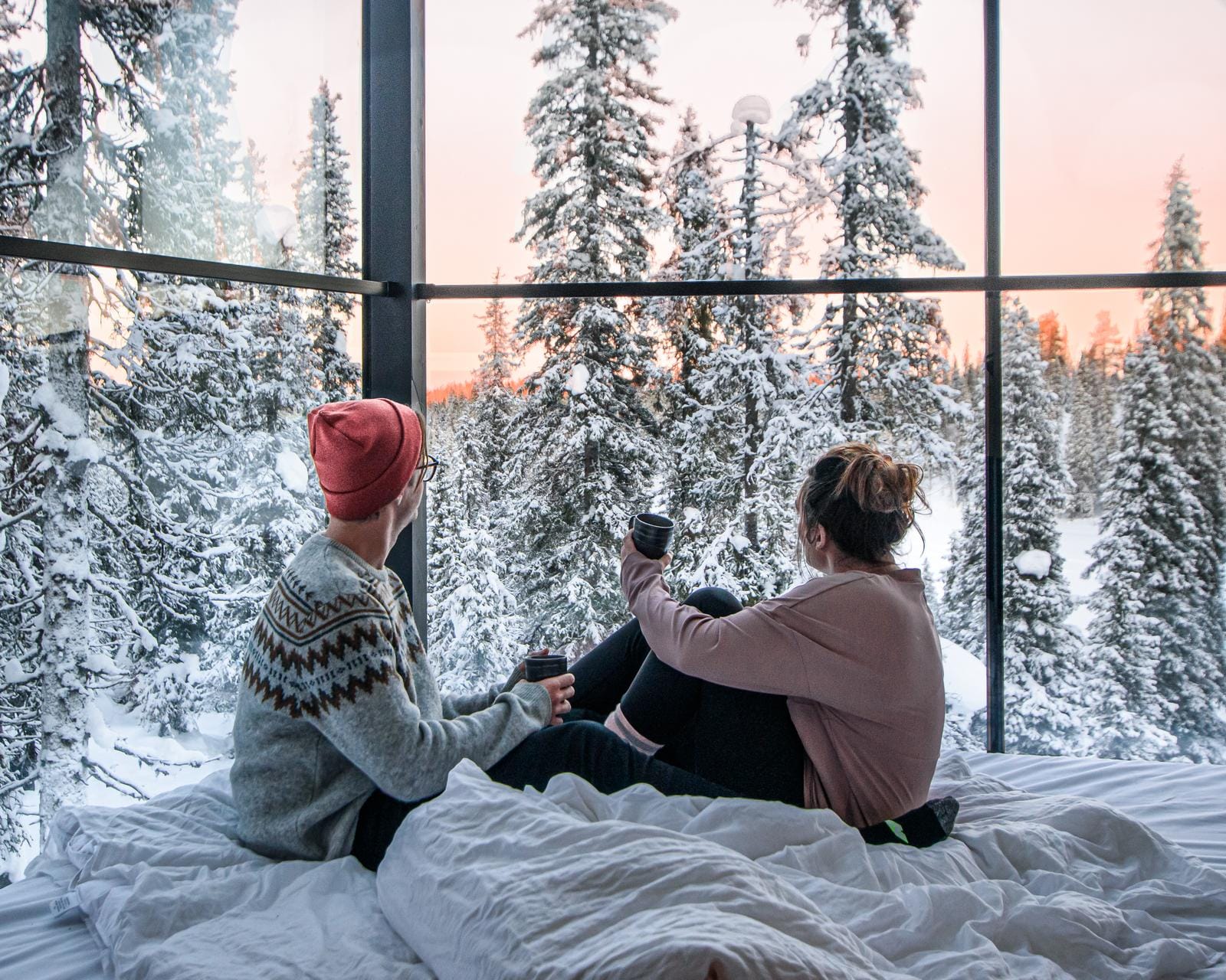 Paar sitzt in verglastem Hotelzimmer mit Blick auf verschneiten Wald bei Sonnenuntergang.
