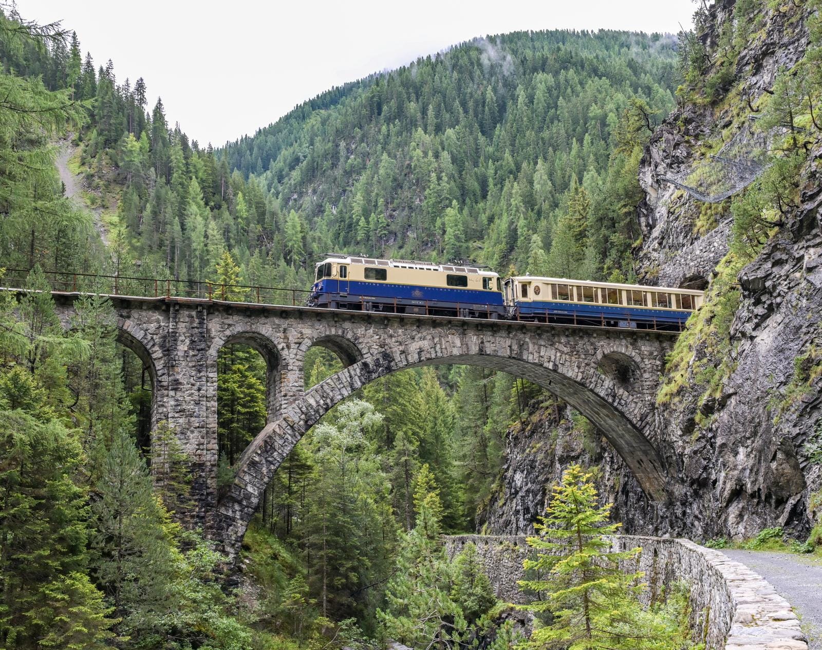 Ein Zug auf einer Steinbrücke in einer grünen, bewaldeten Berglandschaft.
