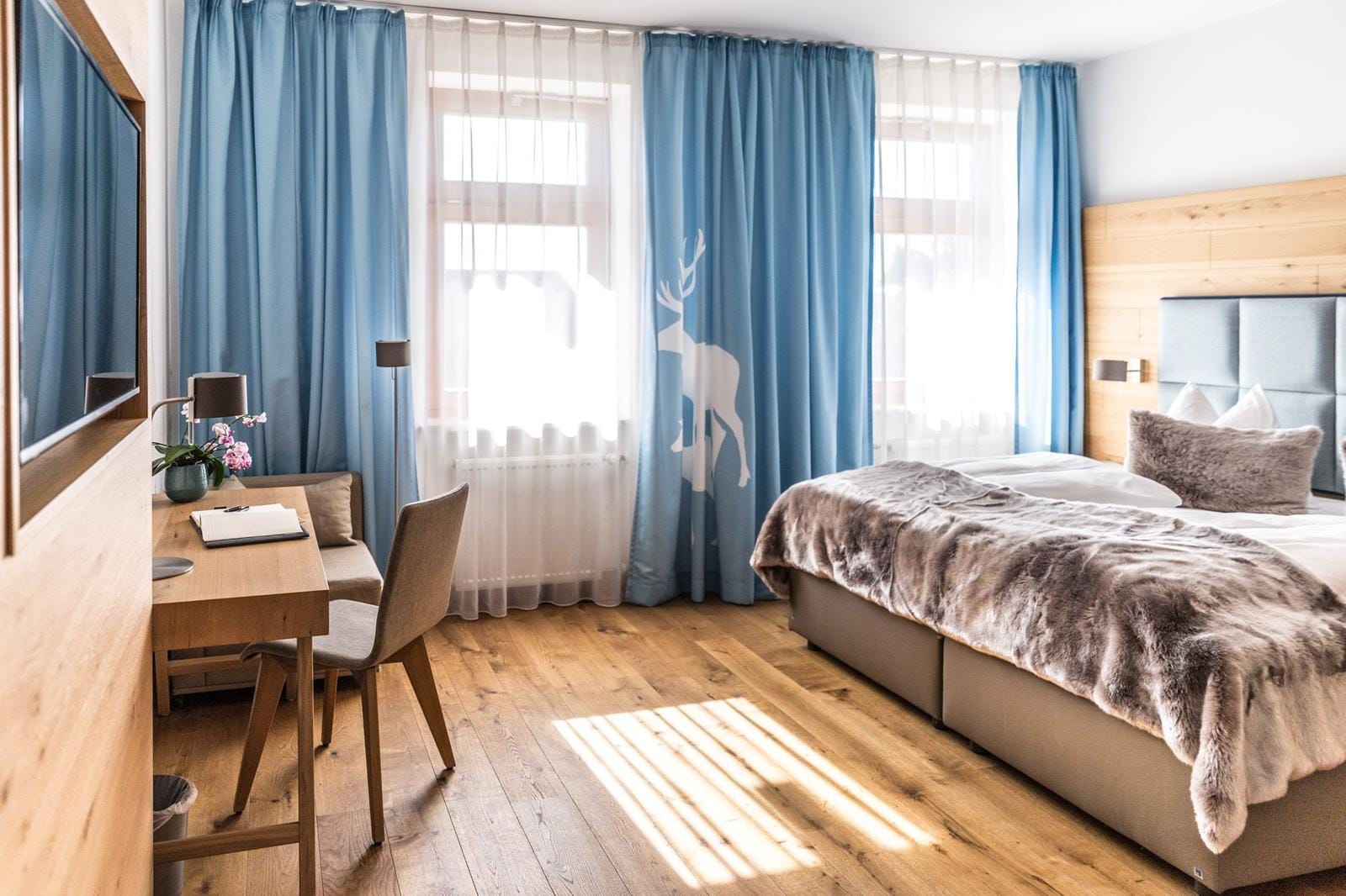 Helles Zimmer mit Holzboden, blauen Vorhängen, gemütlichem Bett und Schreibtisch.