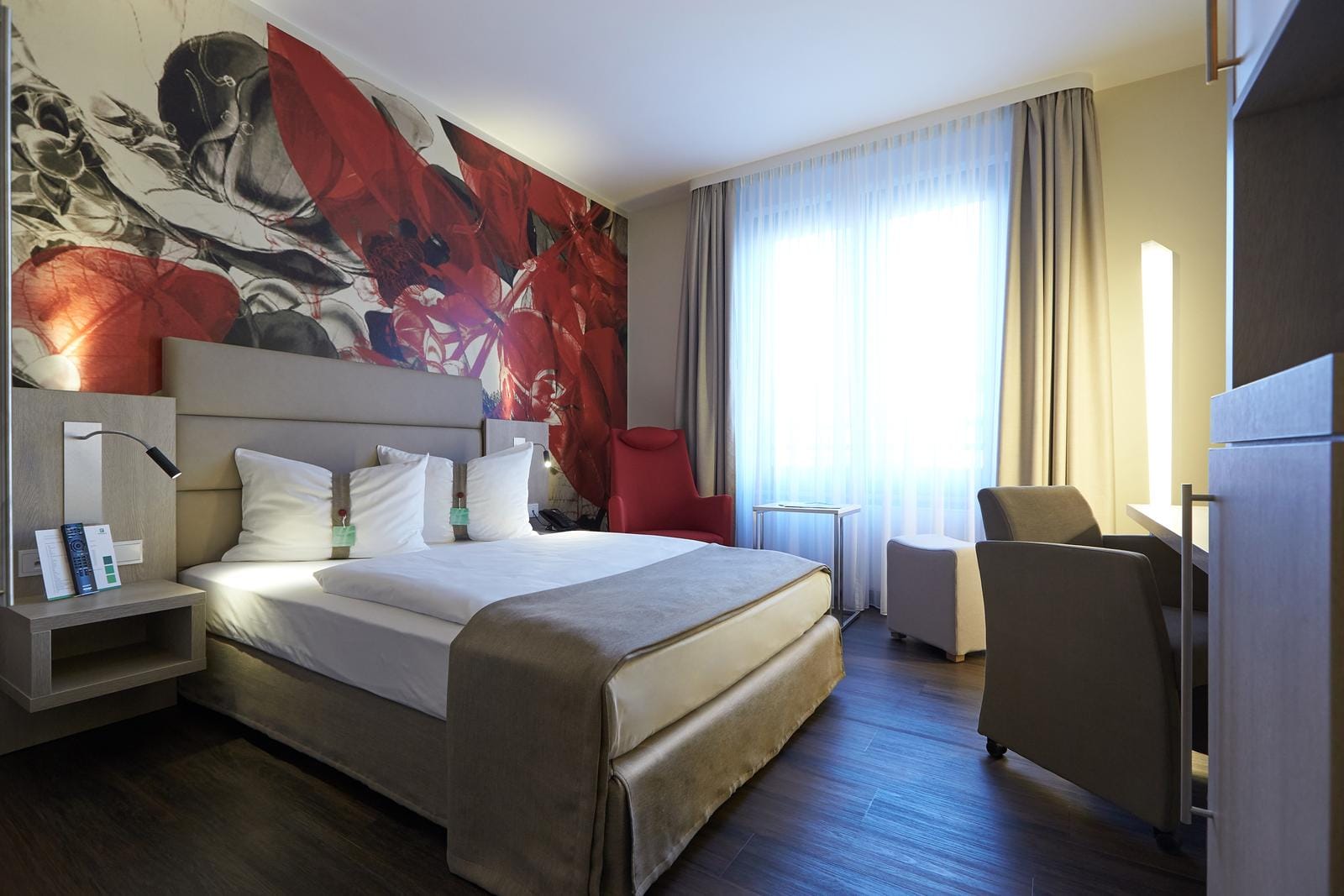 Modernes Hotelzimmer mit Doppelbett, abstrakter Kunst, rotem Sessel und Tageslicht.