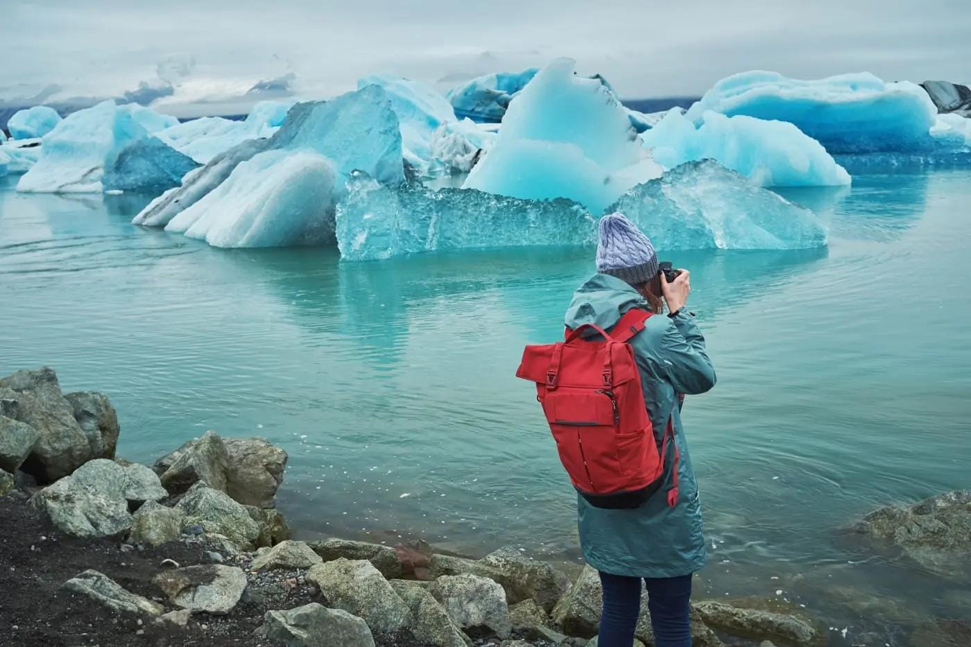 Person mit rotem Rucksack fotografiert blaue Eisberge an einem isländischen See.