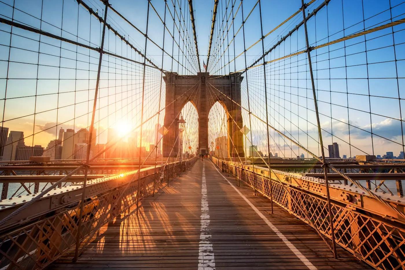 Brooklyn Bridge, New York, bei Sonnenuntergang; orangefarbene Beleuchtung und blauer Himmel.