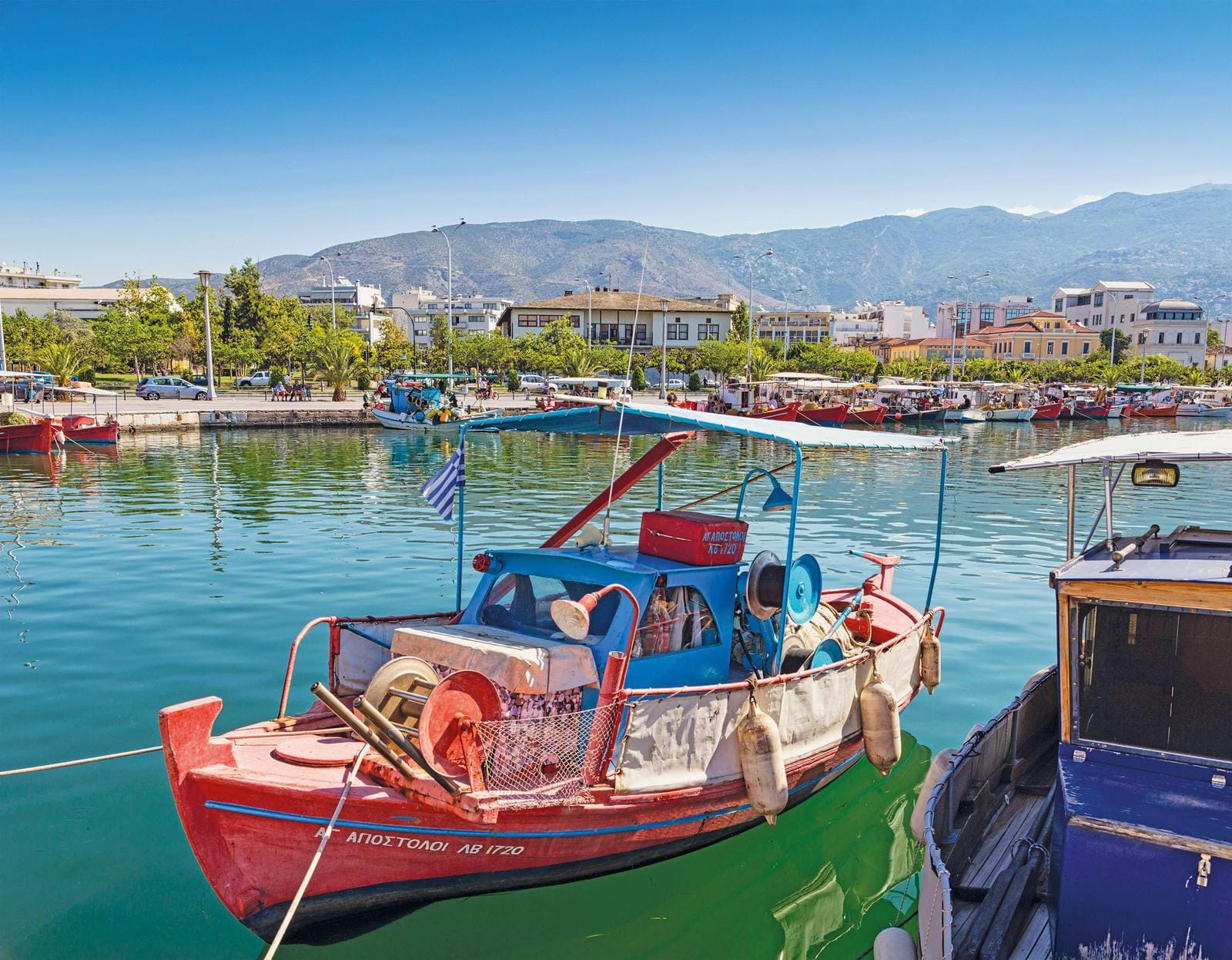 Griechische Hafenstadt mit bunten Booten, Bergen im Hintergrund und blauem Himmel.