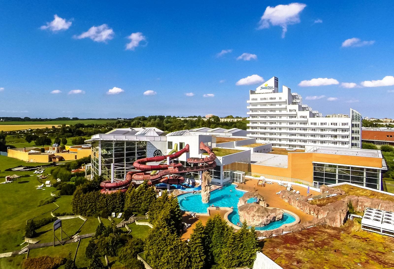 Weißes Hotel mit Wasserrutschen und Außenpool, umgeben von grüner Landschaft.