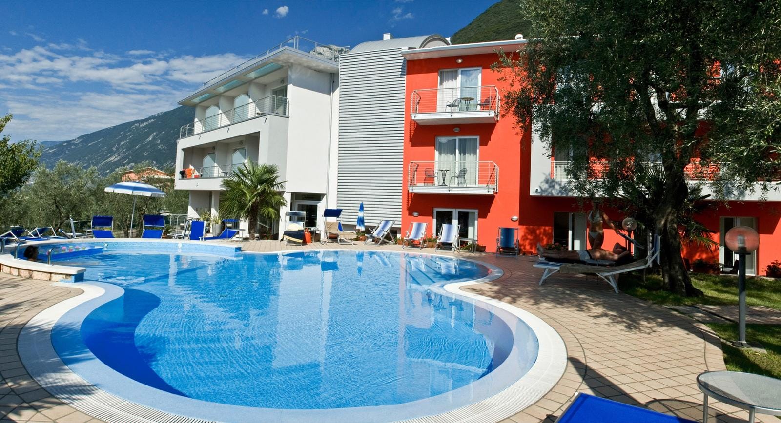 Modernes Hotel mit blauem Pool, Liegestühlen und einem roten Gebäudeflügel.