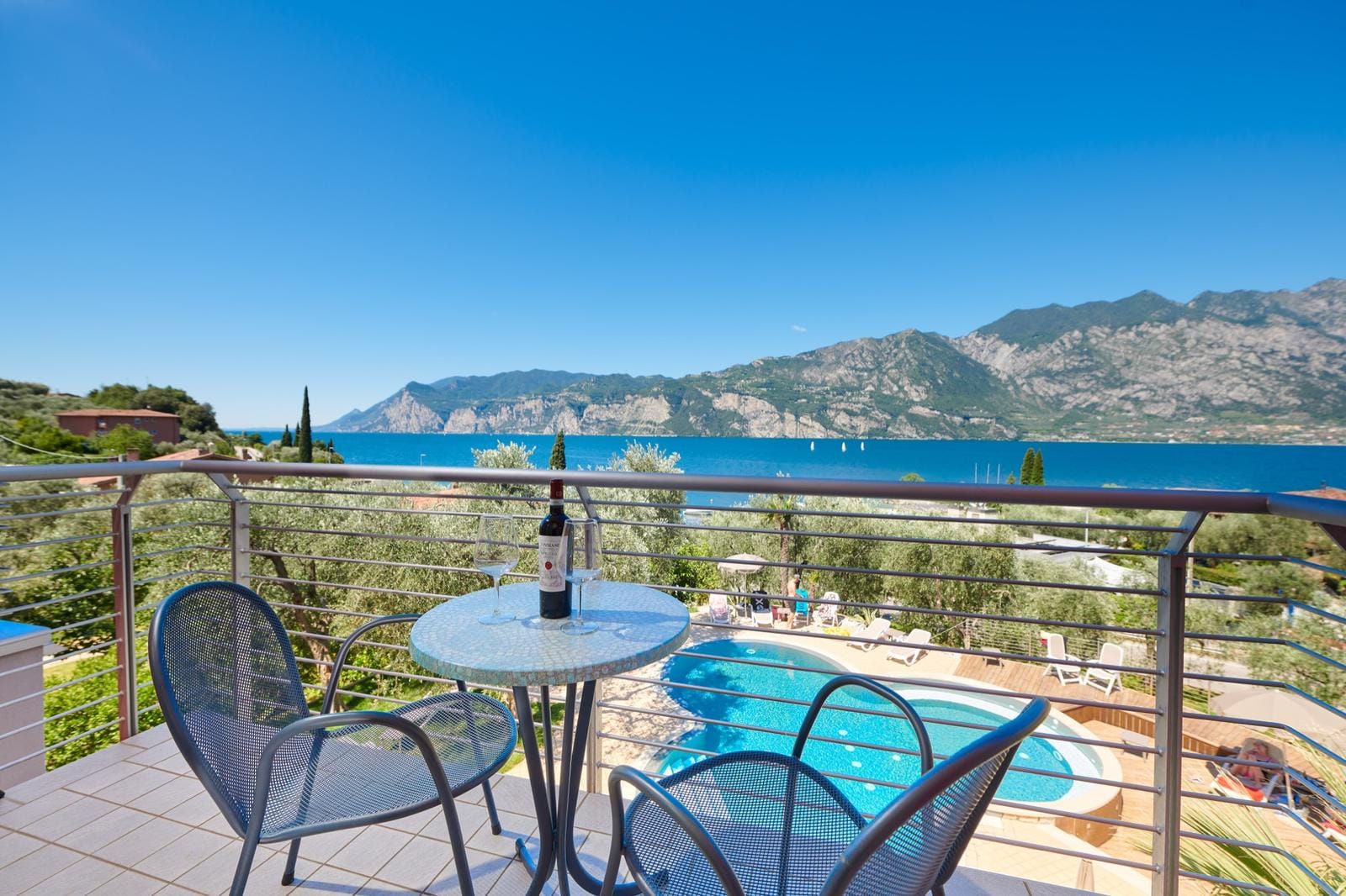 Balkon mit Bergblick am See, runder Tisch, zwei Stühle, Pool und Weinflasche.