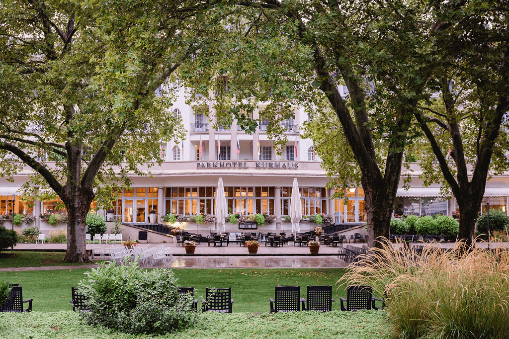 Elegantes Hotel mit klassischer Fassade, Garten und Terrasse unter schattenspendenden Bäumen.