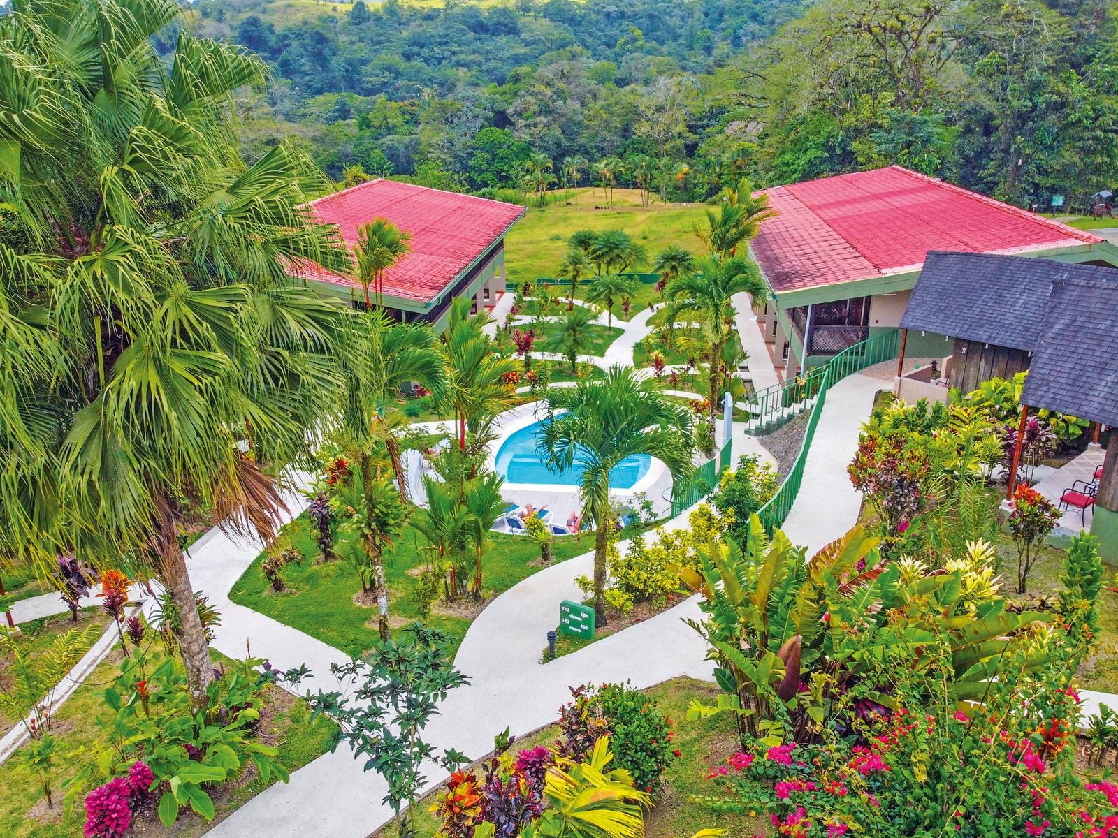 Tropische Hotelanlage mit Pool, Palmen, bunten Blumen und roten Dächern, grüne Umgebung.