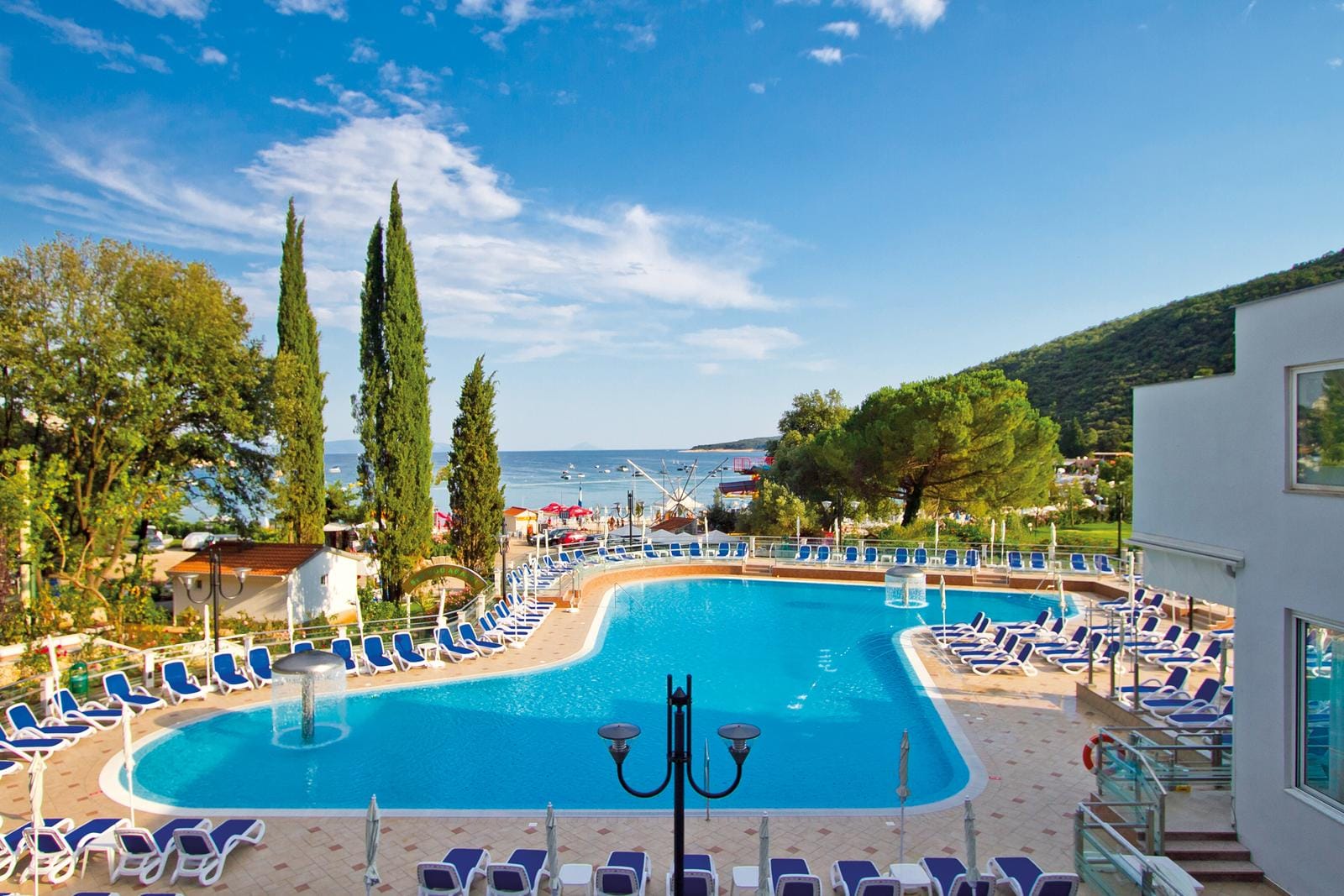 Großer Hotelpool mit blauen Liegen, Meerblick und umgebender grüner Landschaft.