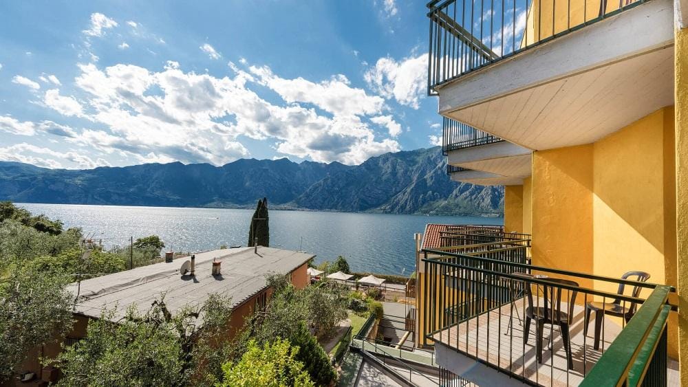 Aussicht auf den Gardasee von einem gelben Balkon mit Bergkulisse und blauem Himmel.