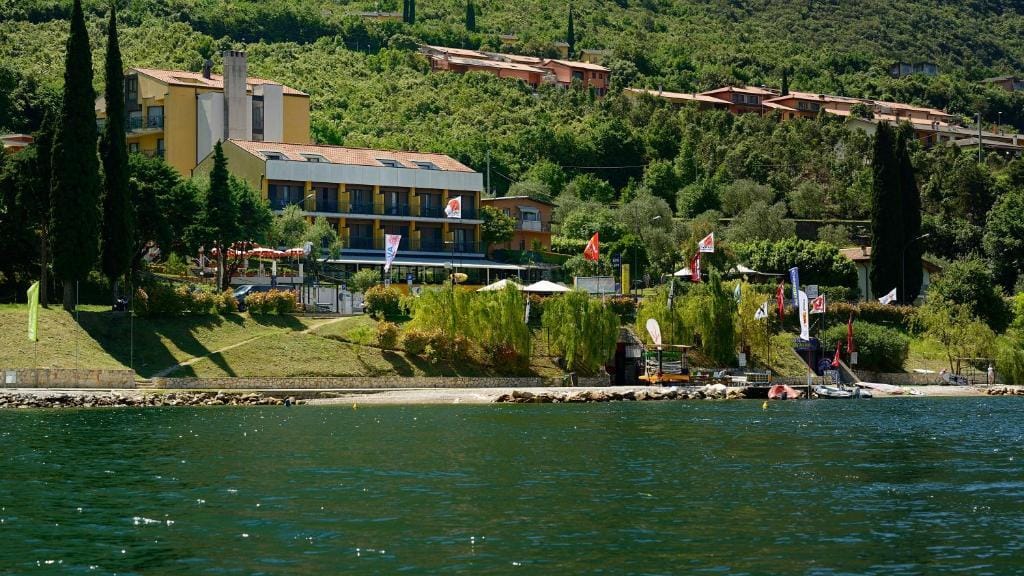 Gelbes Hotel am Seeufer, umgeben von Bäumen und Hügeln, bunte Flaggen am Ufer.