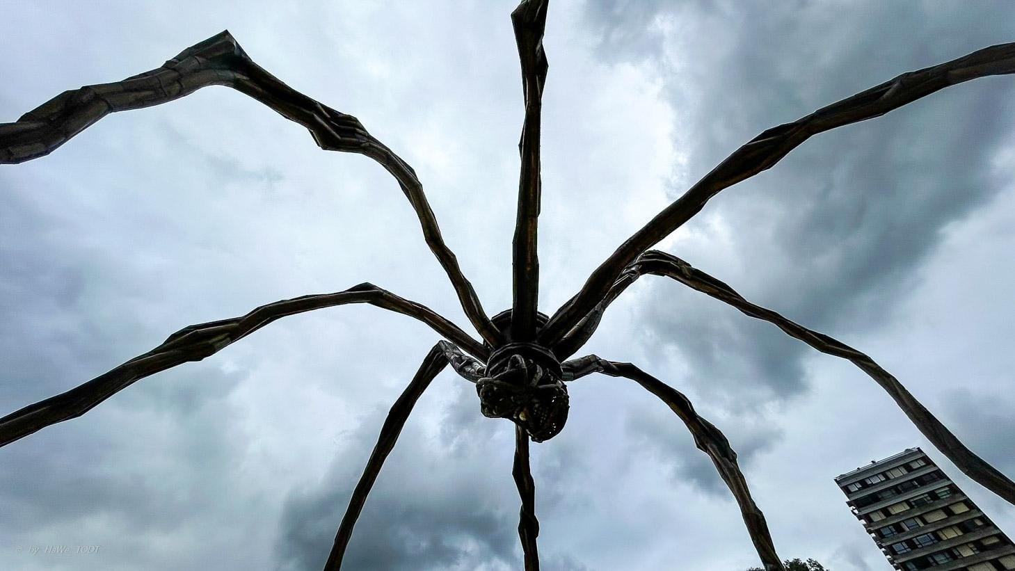 Kunstinstallation große Spinne vor bewölktem Himmel, modernes Gebäude rechts.