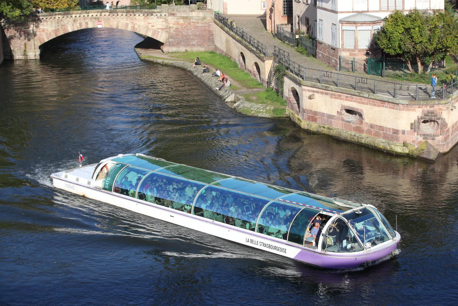 Boot mit Glaskuppel auf Fluss in Straßburg, vorbei an Brücke und steinernem Ufer.
