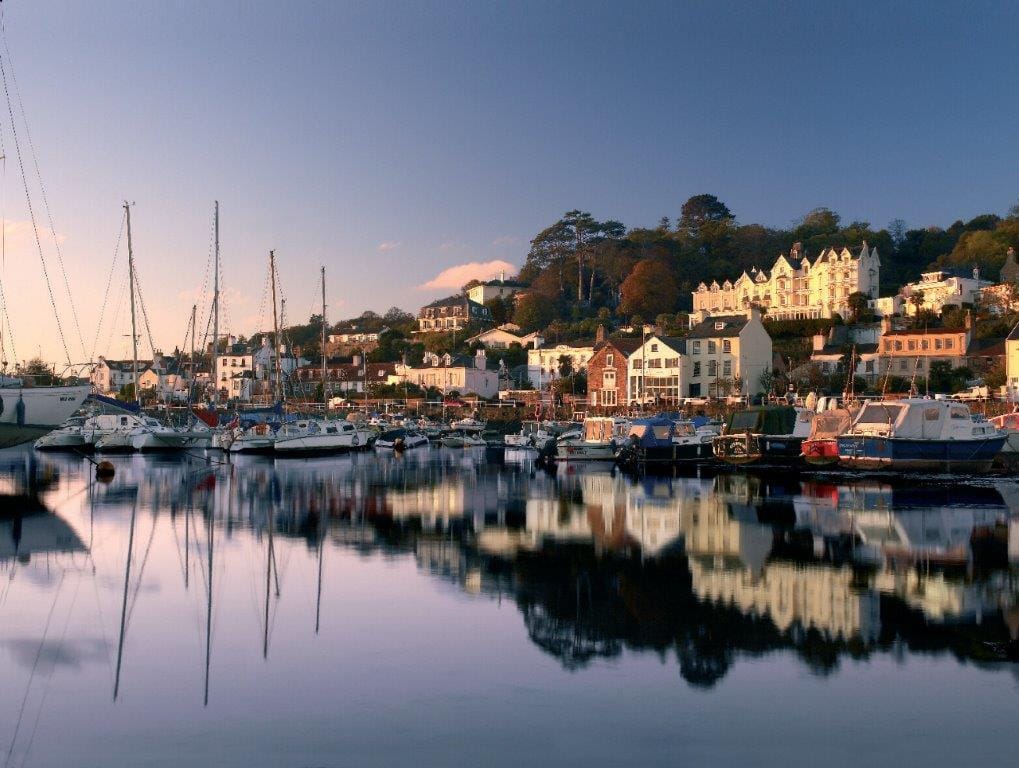 Malebene Hafenstadt mit Segelbooten und Häusern im Sonnenuntergang, ruhiges Wasser.