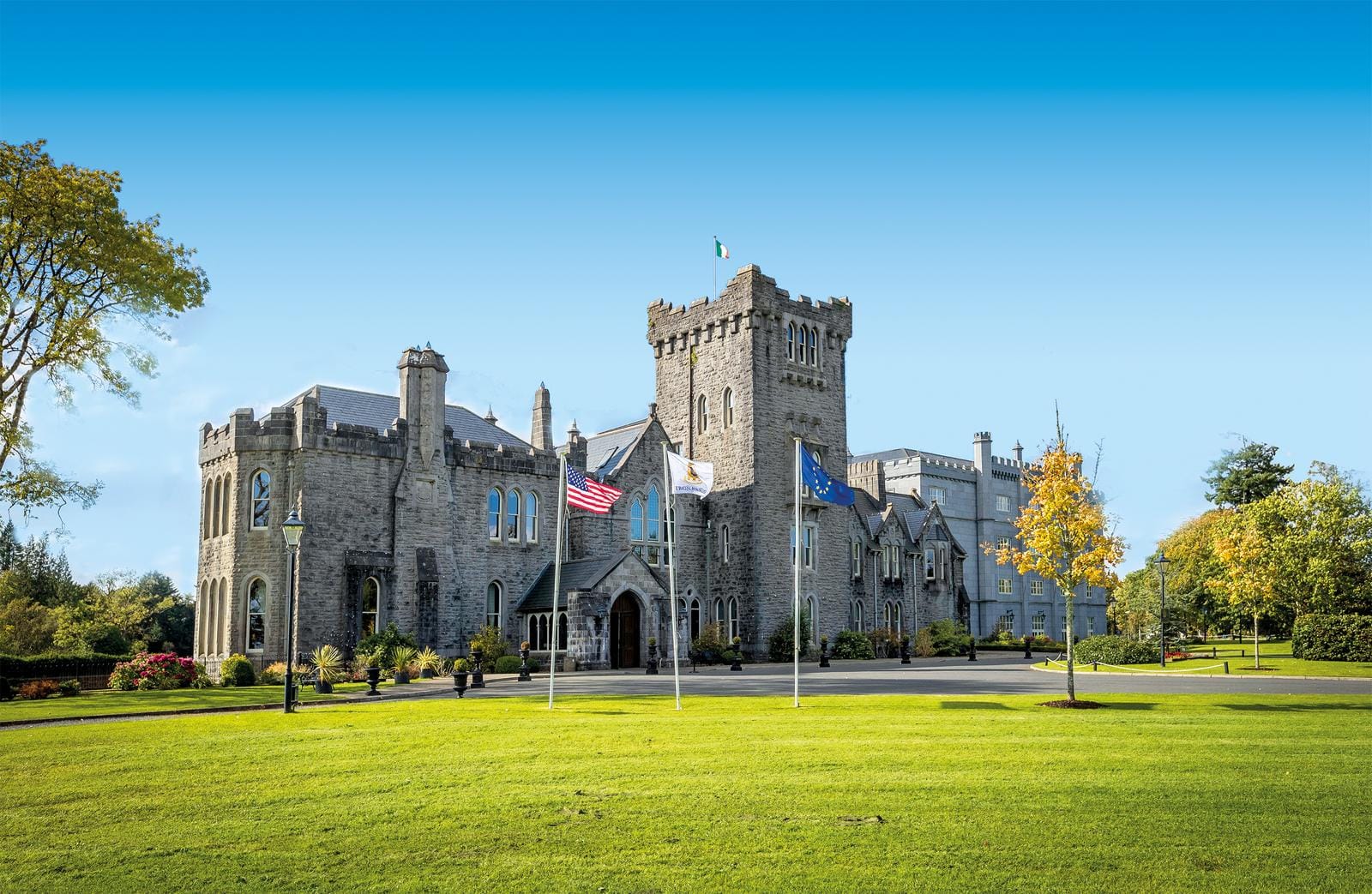 Steinernes Schloss mit Fahnen, grüner Rasen, Herbstbäume, blauer Himmel.