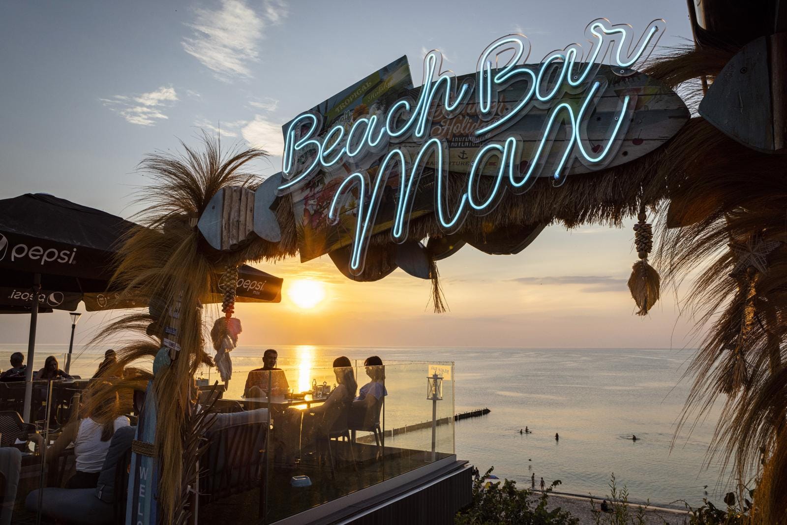 Blick auf eine Strandbar bei Sonnenuntergang, neonbeleuchtetes Schild, entspannte Atmosphäre.